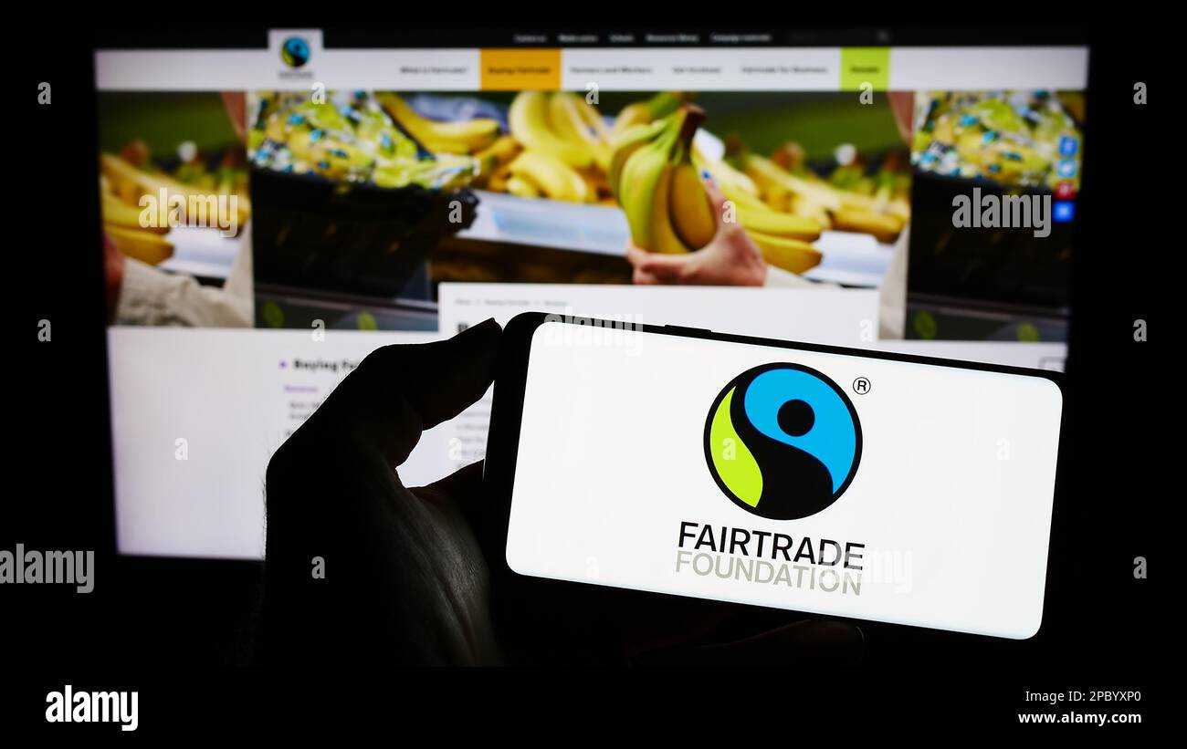 Persona che tiene il telefono cellulare con il logo dell'organizzazione di beneficenza la Fondazione Fairtrade sullo schermo davanti alla pagina web. Messa a fuoco sul display del telefono. Foto Stock