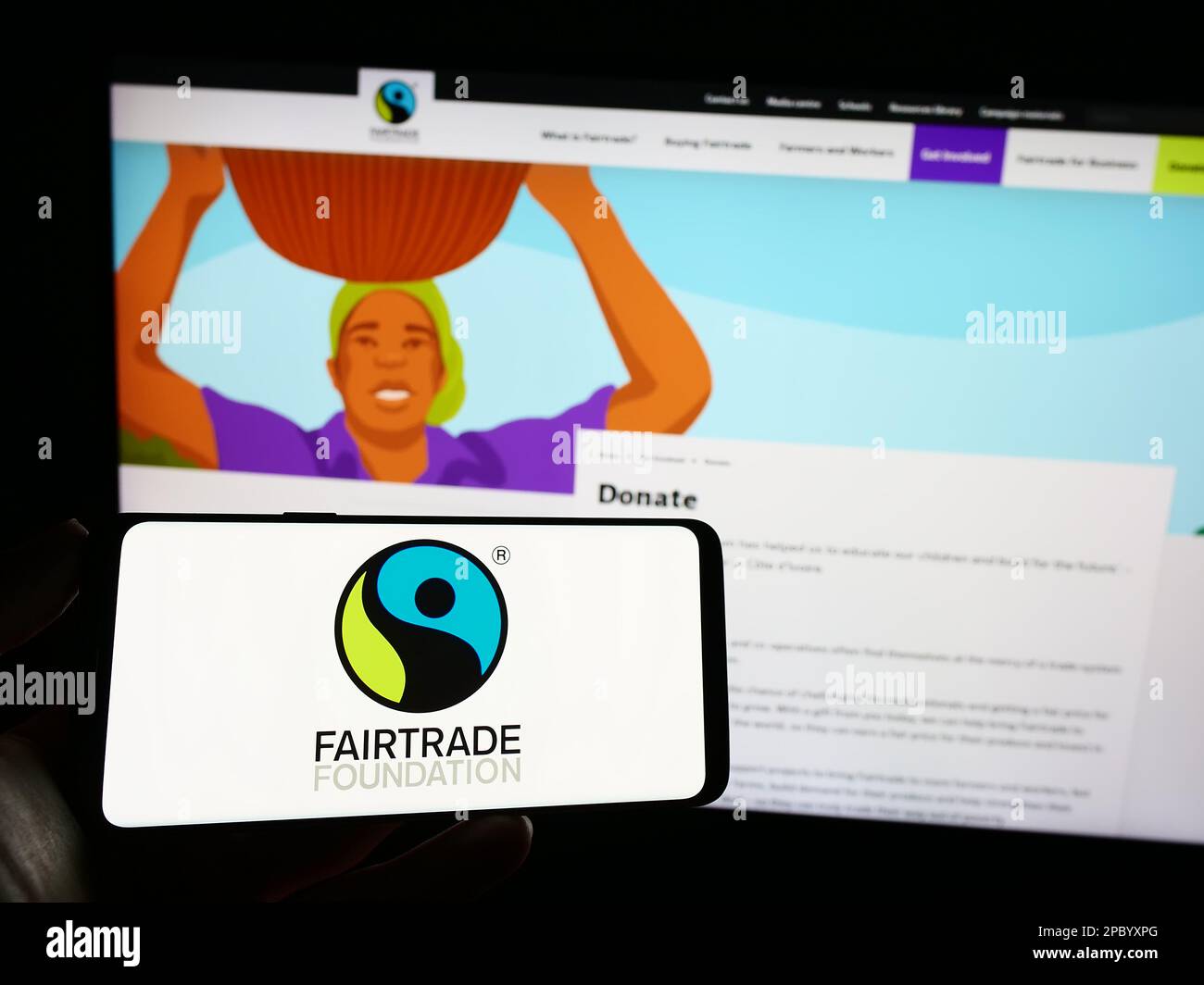 Persona che tiene il cellulare con il logo dell'organizzazione di beneficenza la Fondazione Fairtrade sullo schermo di fronte alla pagina web. Messa a fuoco sul display del telefono. Foto Stock