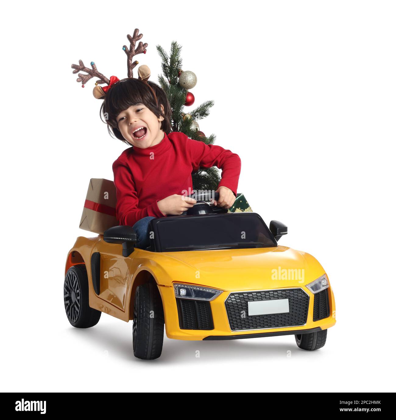 Carino ragazzino con albero di Natale e scatola regalo che guida l'auto giocattolo elettrica dei bambini su sfondo bianco Foto Stock