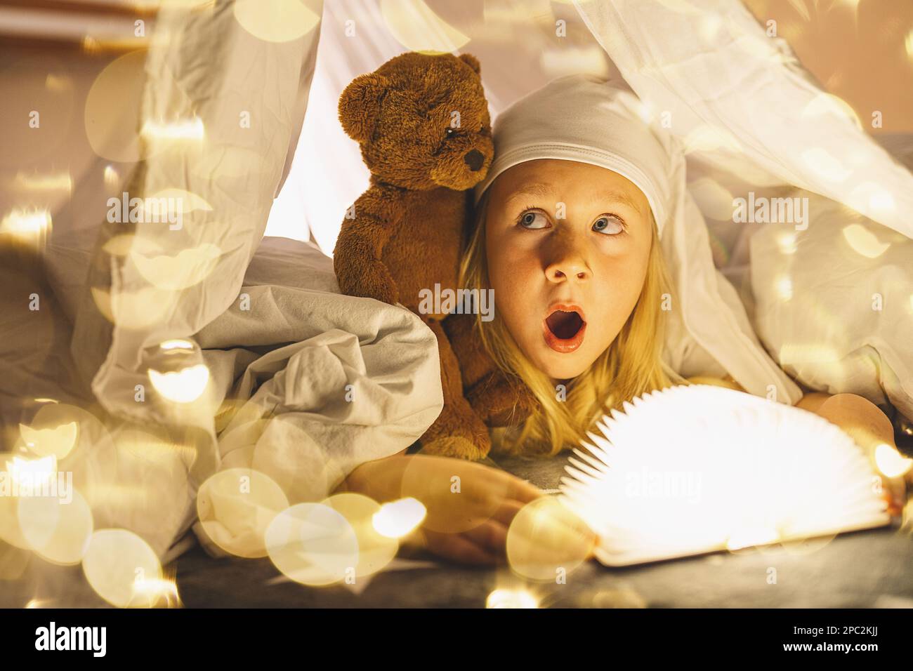 bambina che legge il libro con orsacchiotto sotto coperta di notte, prima di andare a letto, luce magica che esce dal libro. Foto Stock