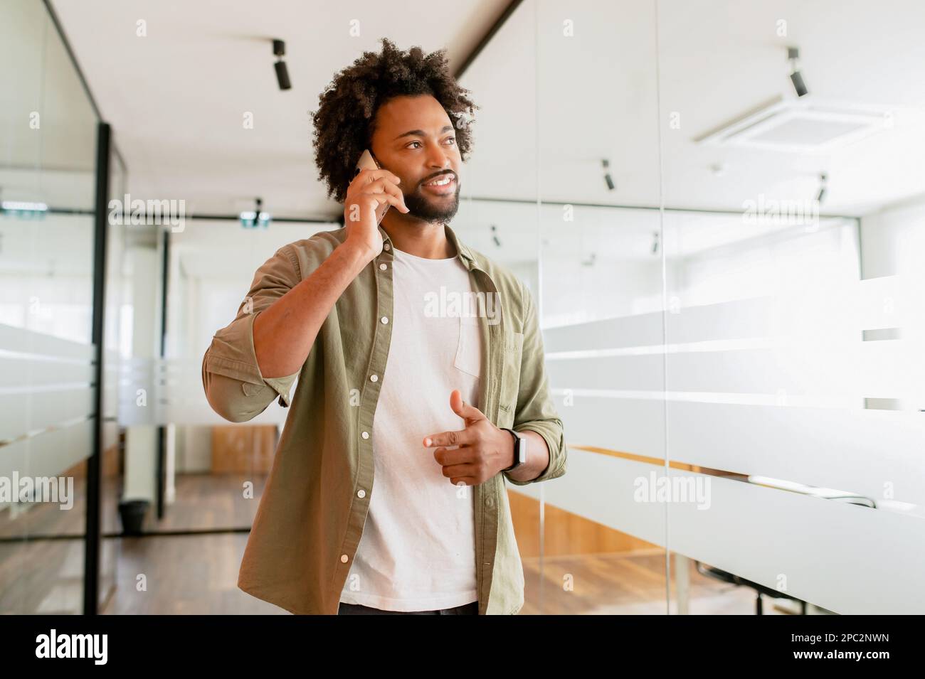 Dipendente di un ufficio di sesso maschile che ha una conversazione telefonica, in contatto con i clienti. Uomo afro-americano sicuro in abbigliamento casual elegante parlare sullo smartphone mentre cammina in ufficio Foto Stock