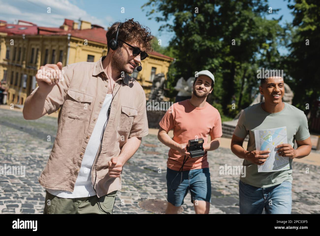 Guida positiva in occhiali da sole e cuffie leader tour per i turisti multietnici con mappa e fotocamera d'epoca sulla discesa Andrews a Kiev, immagine stock Foto Stock