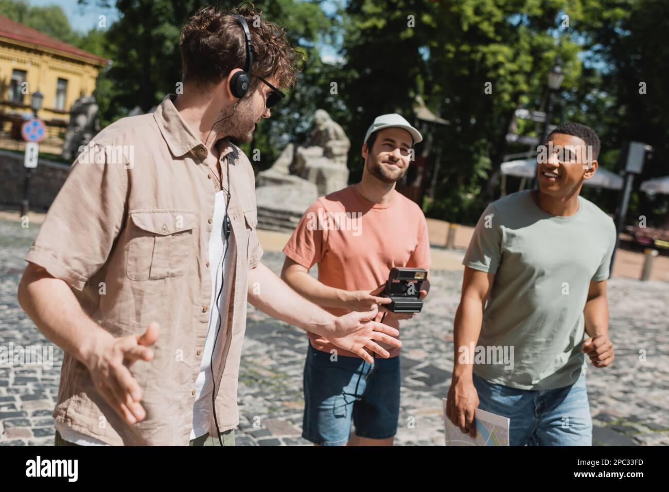 Guida turistica in cuffie gesturing vicino a turisti multietnici a piedi con mappa e fotocamera d'epoca sulla discesa Andrews a Kiev, immagine stock Foto Stock