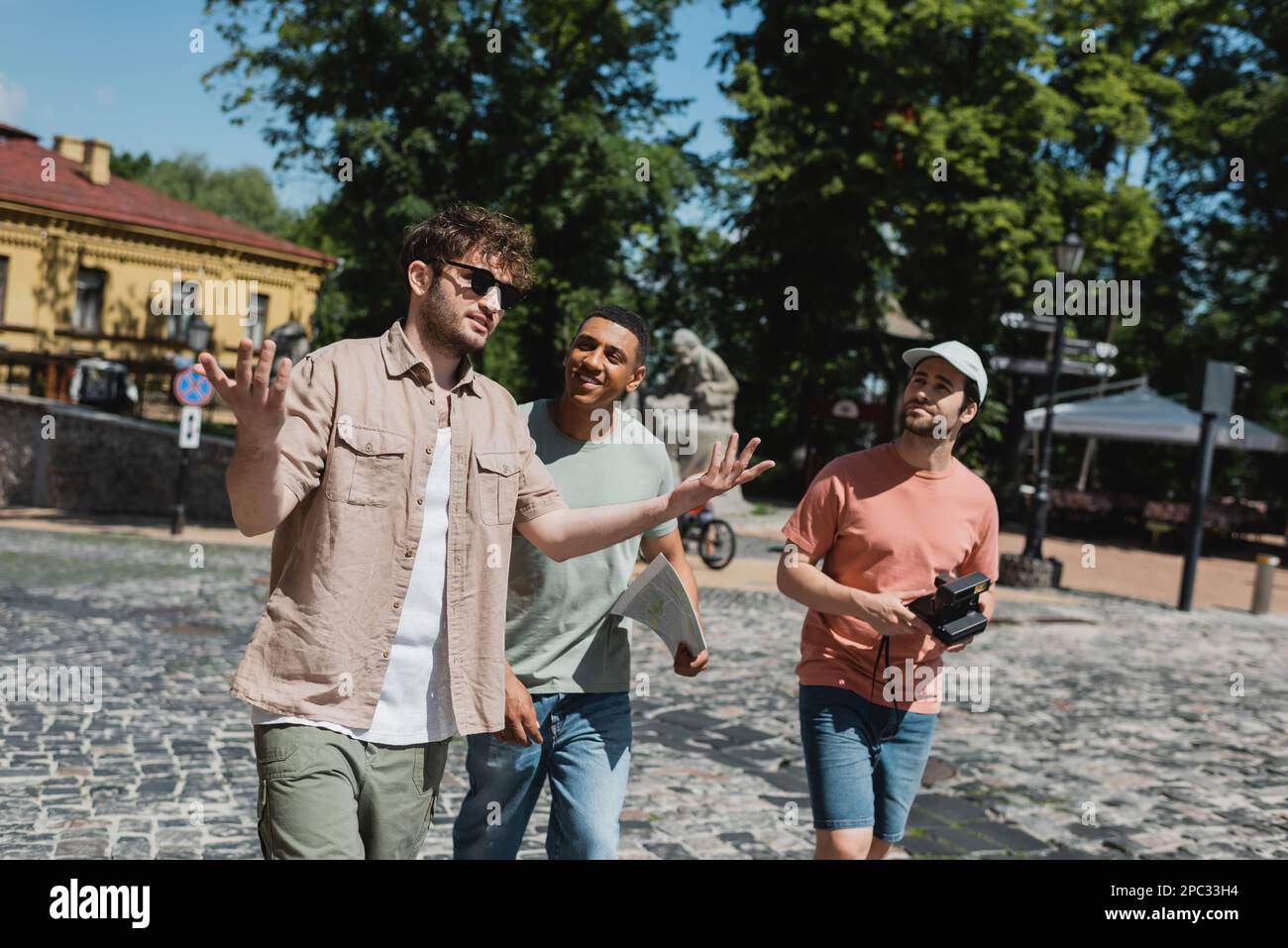 Guida del tour in occhiali da sole gesturing mentre si parla con i turisti multiculturali durante la passeggiata sulla discesa Andrews a Kiev, immagine stock Foto Stock