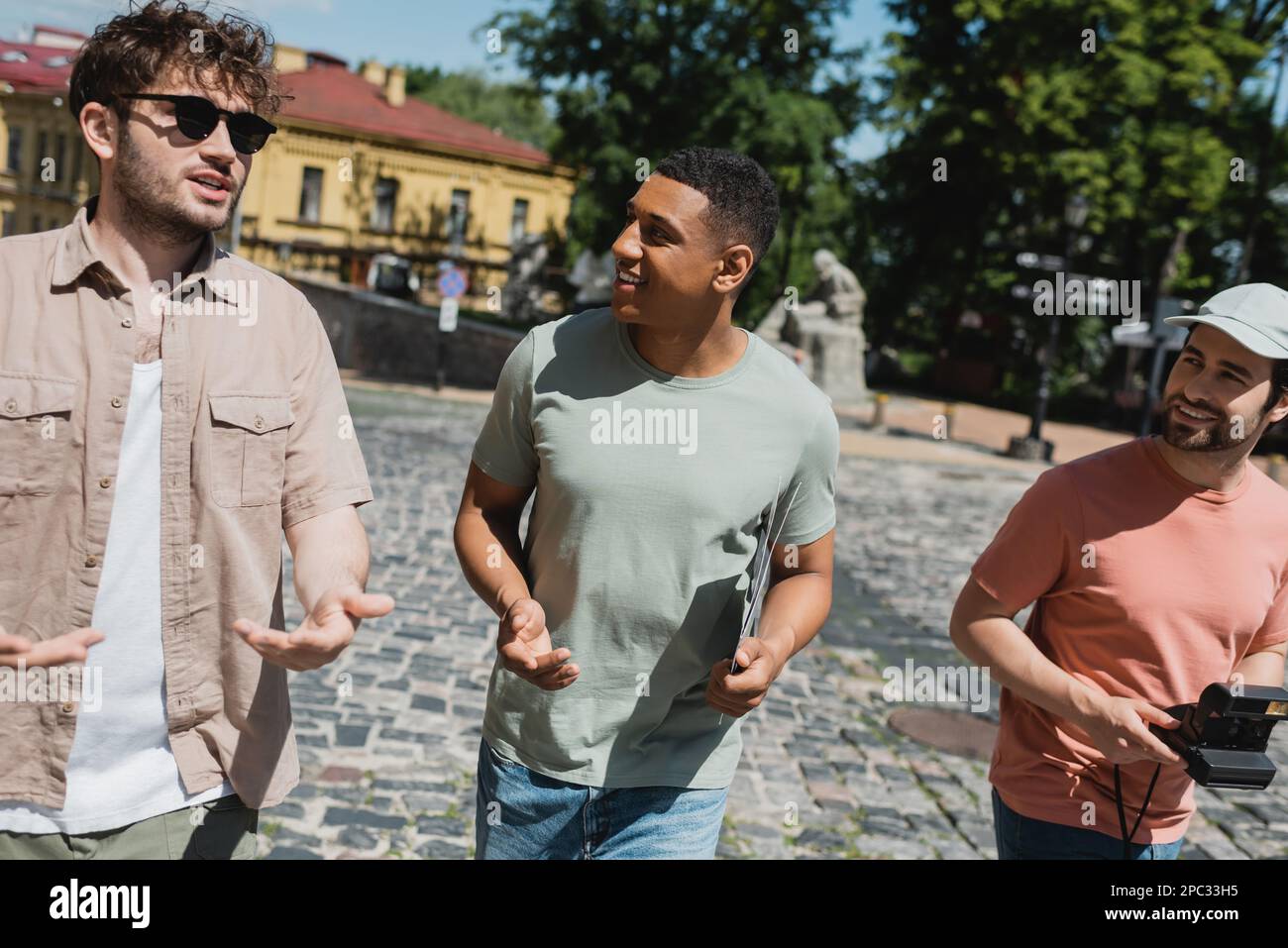 Sorridenti uomini multietnici che ascoltano una giovane guida in occhiali da sole mentre camminano sulla discesa di Andrews a Kyiv, immagine di scorta Foto Stock