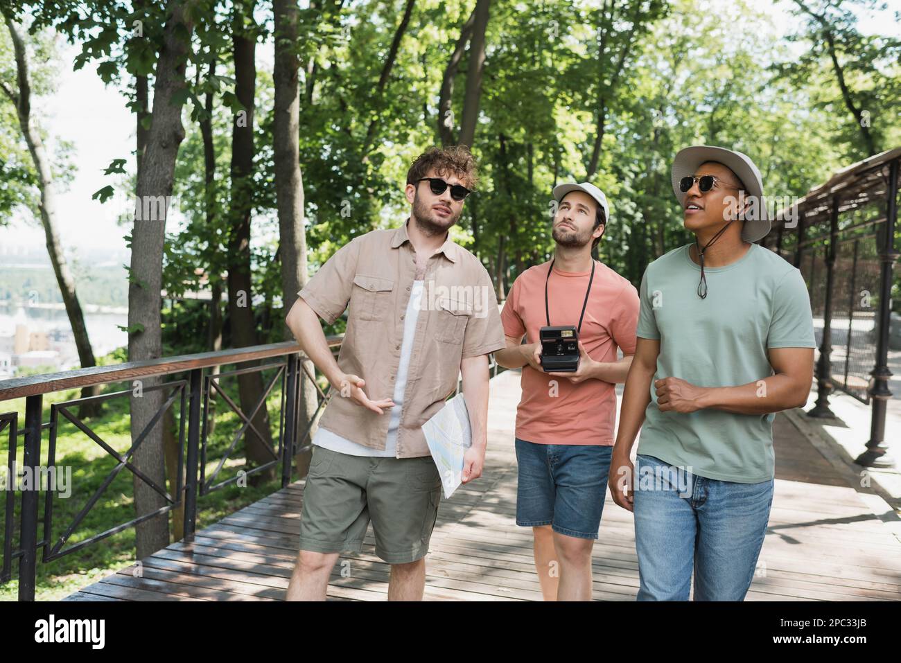 viaggiatori multiculturali in cappelli di sole che guardano via durante la passeggiata estiva con guida turistica nel parco urbano, immagine stock Foto Stock