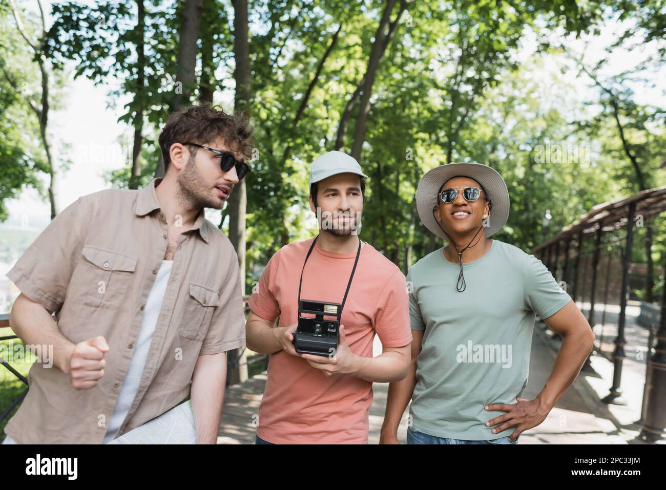 giovane guida turistica in occhiali da sole che parla con i turisti multietnici in cappelli di sole durante l'escursione nel parco urbano, immagine stock Foto Stock
