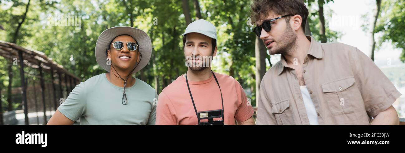 giovane guida turistica in occhiali da sole che parla con i turisti multietnici indossando cappelli da sole nel parco verde, banner, immagine stock Foto Stock