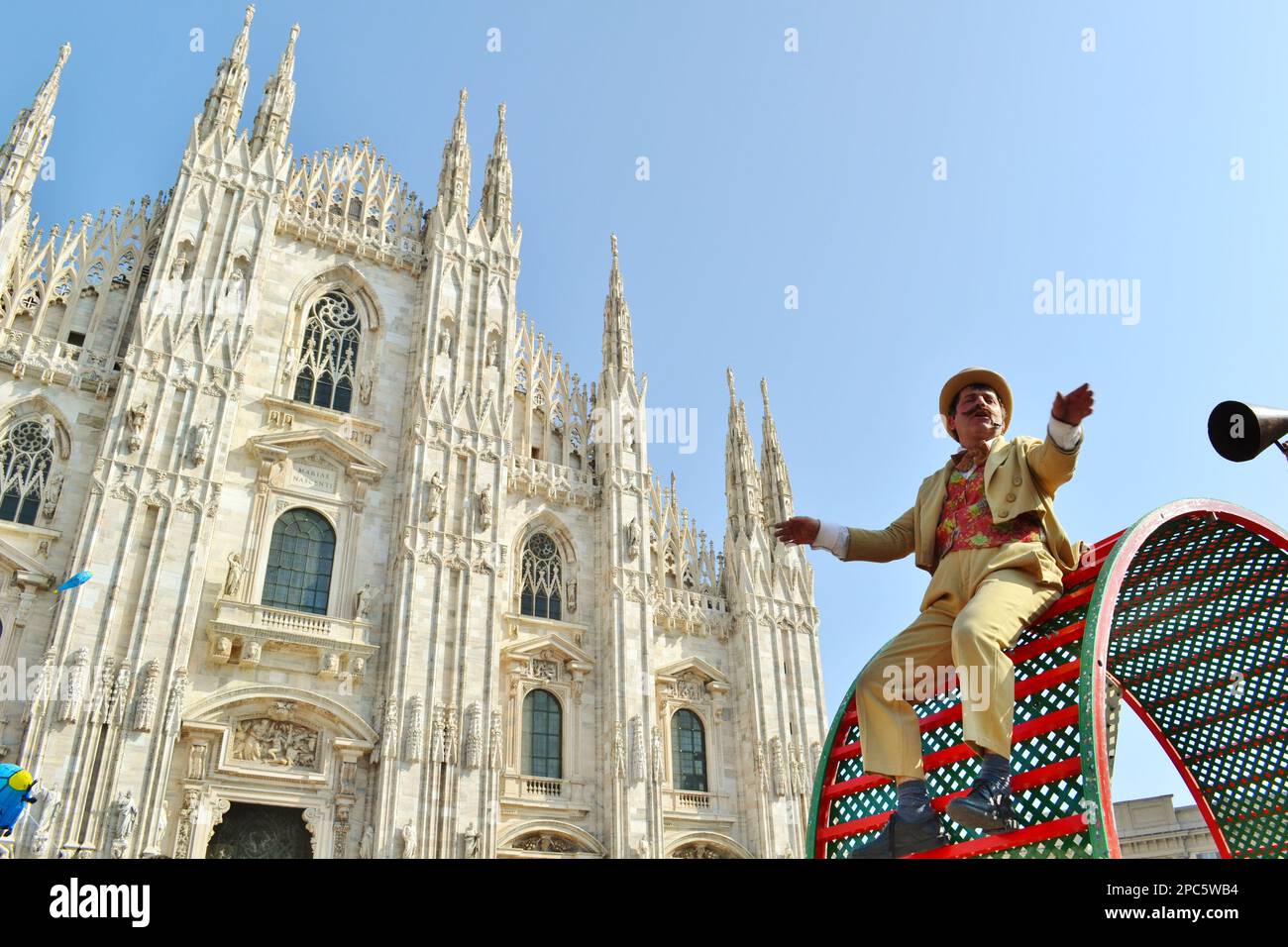 L'esecutore del circo vestito in una maschera di carnevale del cilindro del cappello sta cadendo giù la ruota del criceto. Sullo sfondo il Duomo di Milano. Foto Stock