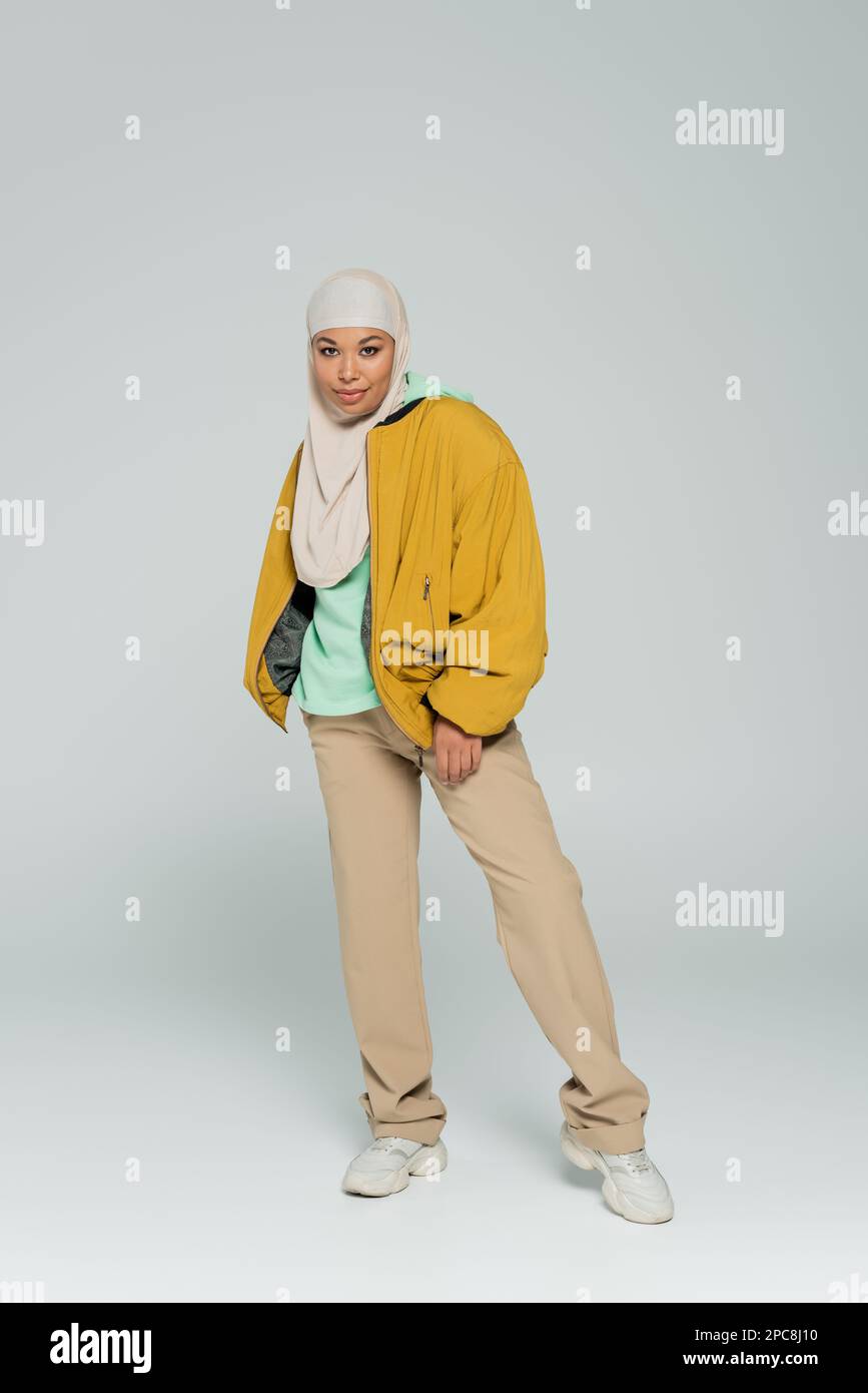 intera lunghezza di donna multirazziale in hijab e abbigliamento casual alla moda in piedi e sorridente alla macchina fotografica su sfondo grigio, immagine stock Foto Stock