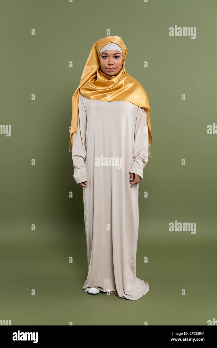 intera lunghezza di donna musulmana multirazziale in hijab di seta gialla e abito abaya in piedi su sfondo verde oliva, immagine stock Foto Stock