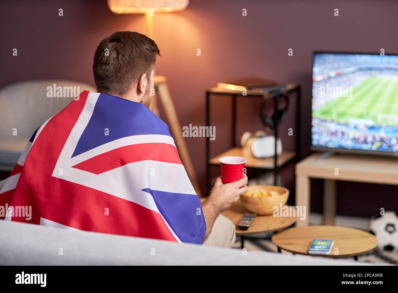 Vista posteriore del tifoso sportivo che indossa la bandiera del Regno Unito mentre guarda la partita di calcio a casa, spazio copia Foto Stock