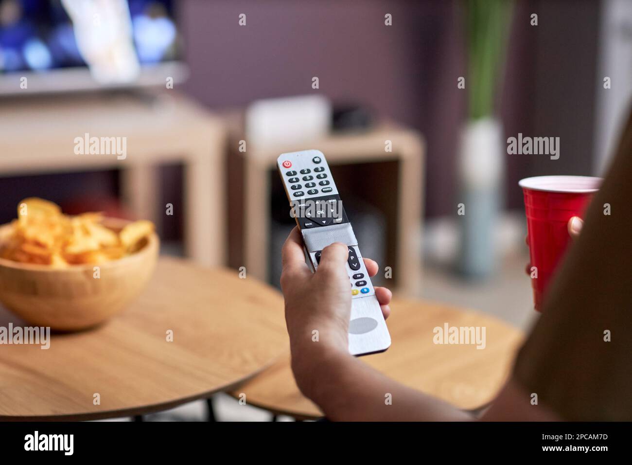 Primo piano della mano maschile con telecomando mentre si guarda la TV a casa e si gustano spuntini, spazio copia Foto Stock