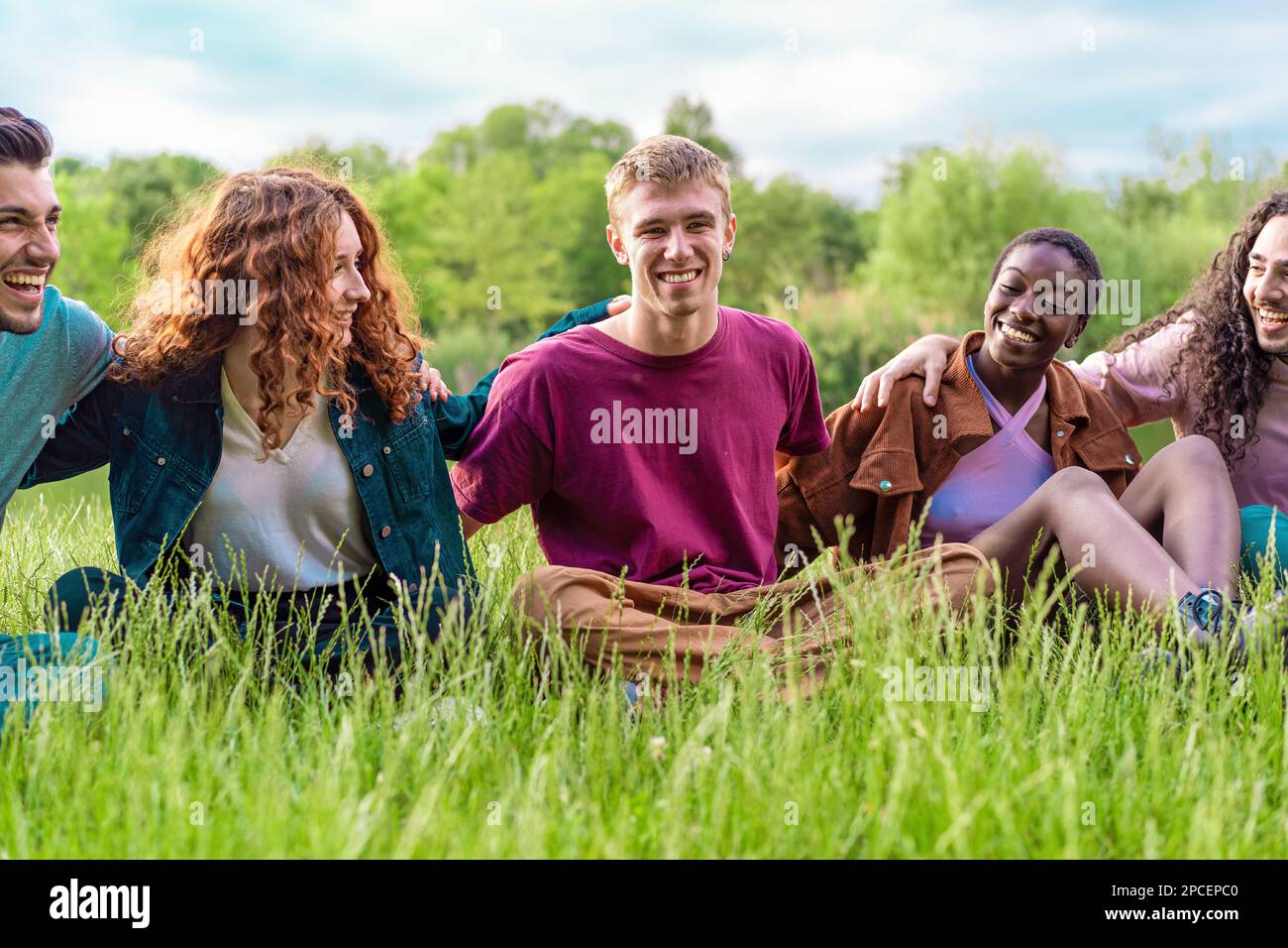 Un gruppo diversificato di amici abbraccia e sorride felicemente mentre si siede sull'erba in un parco - vita all'aperto, amicizia, integrazione razziale, fiducia e ca Foto Stock