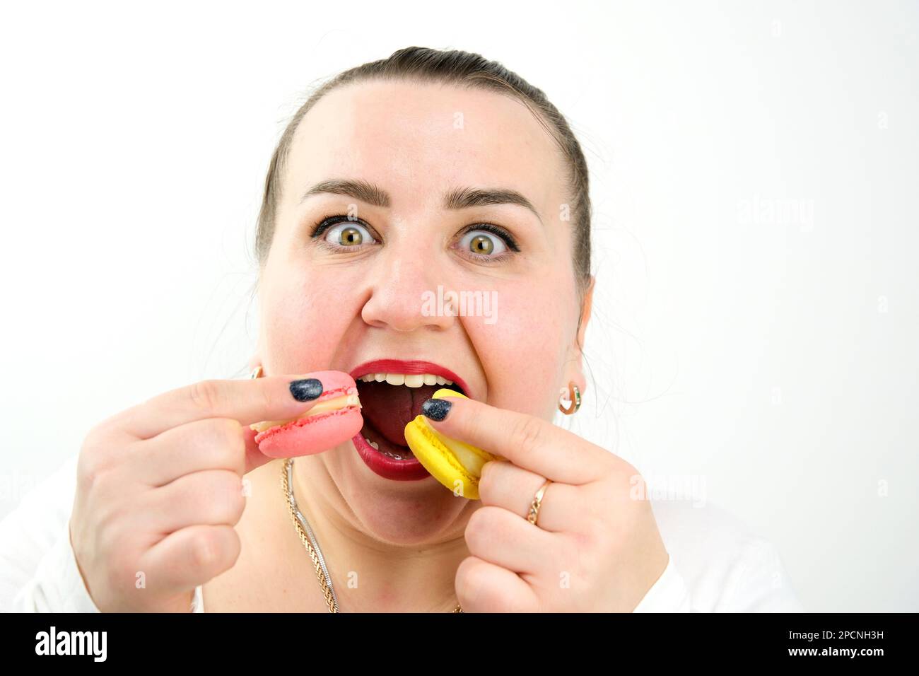 una donna grassa mangia un sacco di deliziosi dolci macarons dolci sorride gioiosamente e bocca aperta larga, gioielli d'oro su uno sfondo bianco, dieta, overeating Foto Stock