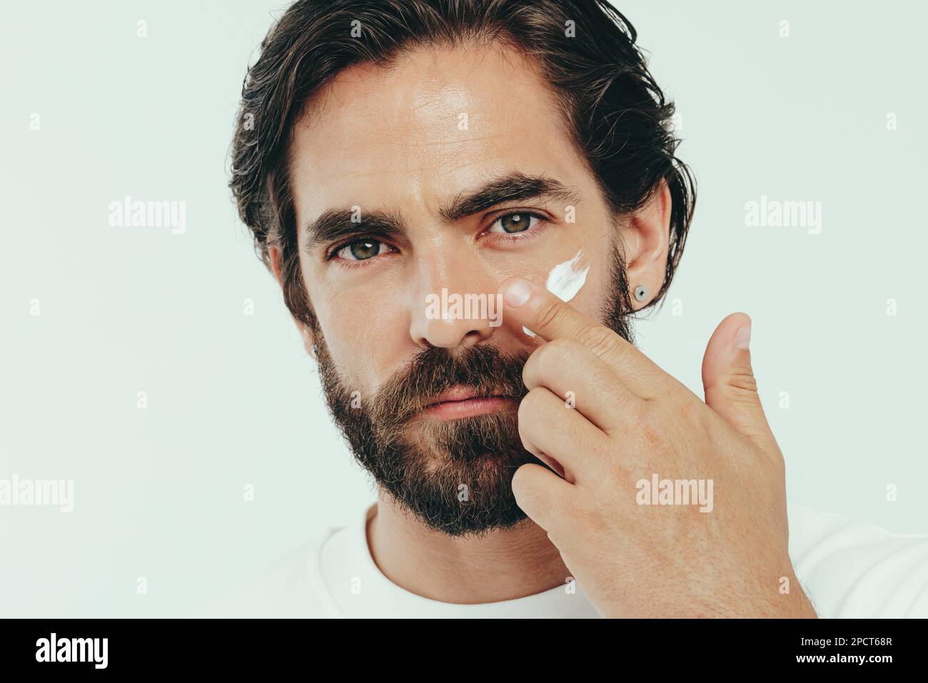 Uomo che guarda la macchina fotografica mentre prende un momento per coccolarsi con una certa cura della pelle di qualità. Uomo sicuro che applica una crema per il viso per mantenere la sua pelle h Foto Stock