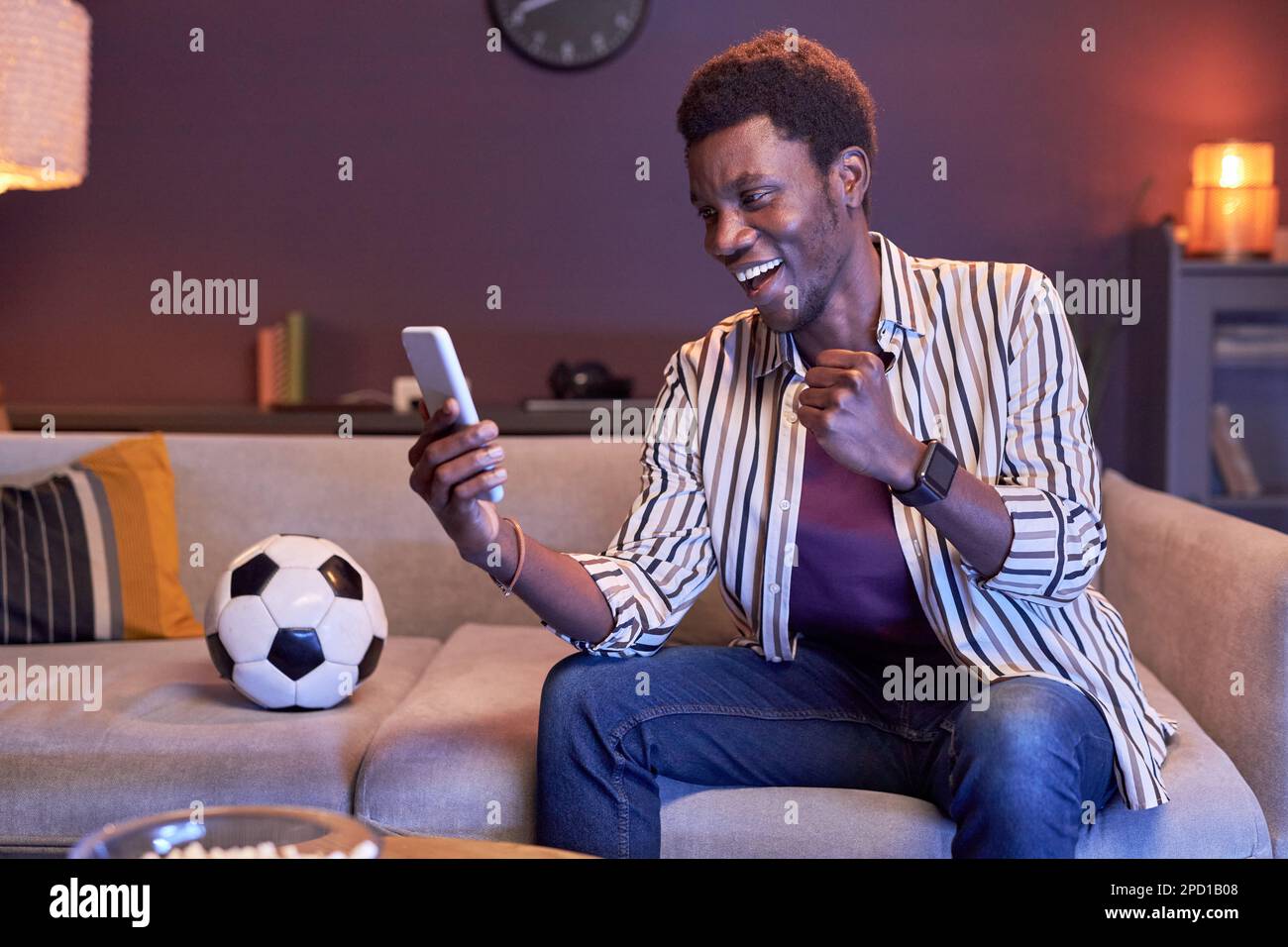 Ritratto di un uomo nero entusiasta che guarda la partita sportiva in TV a casa e utilizzando l'app di scommesse online in luce blu, copia spazio Foto Stock
