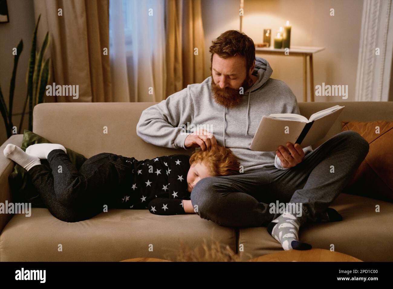 Padre reding libro mentre il suo piccolo figlio carino che dorme sulle sue ginocchia in salotto accogliente a casa Foto Stock