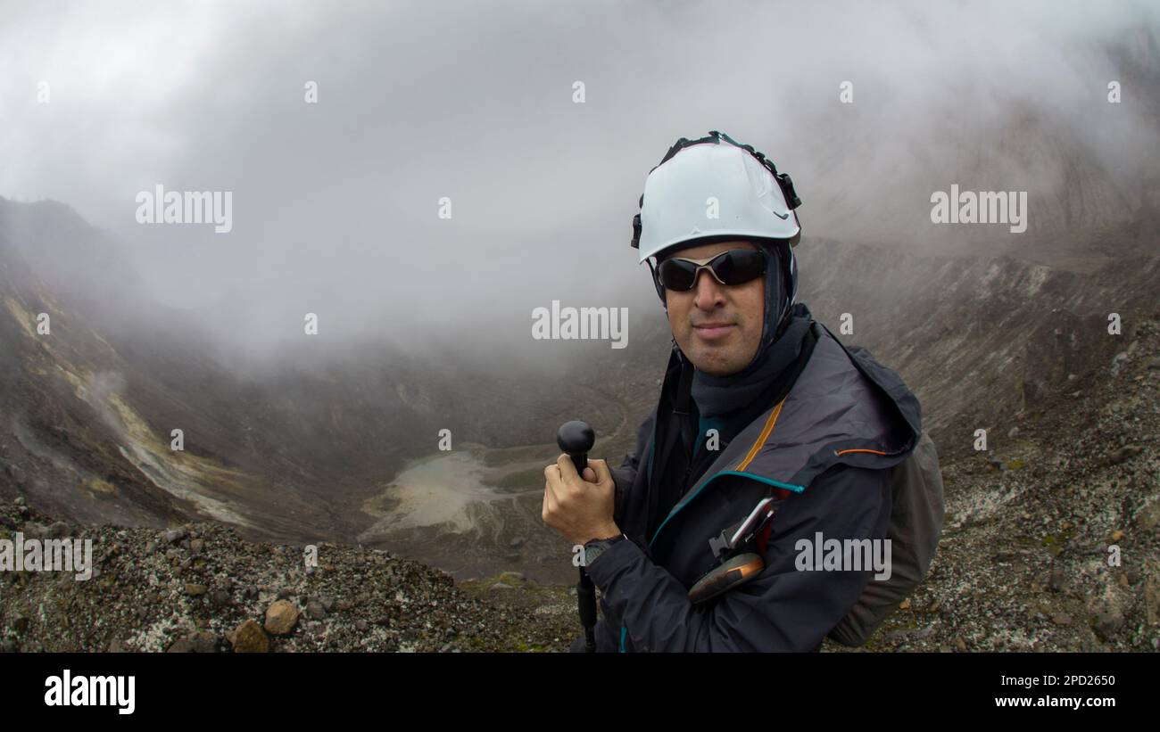 Giovane uomo arrampicatore che cammina con cappotto nero e casco utilizzando il palo da trekking da solo presso il cratere del vulcano Guagua Pichincha in una giornata nuvolosa Foto Stock