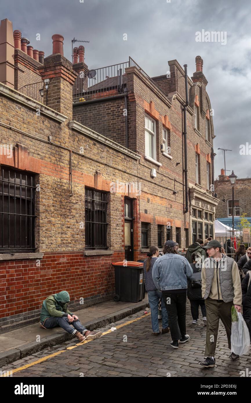 La gente cammina davanti ad una persona vulnerabile che siede da sola sul marciapiede in Ezra Street, Bethnal Green, vicino al popolare mercato dei fiori di Columbia Road W. Foto Stock