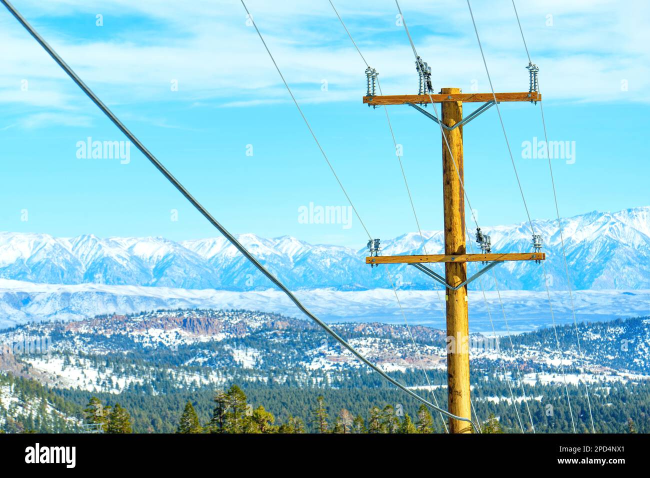 Palo di potenza in legno con fili contro le cime innevate di una catena montuosa e un cielo blu chiaro, sottolineando l'importanza di un affidabile sostegno elettrico Foto Stock
