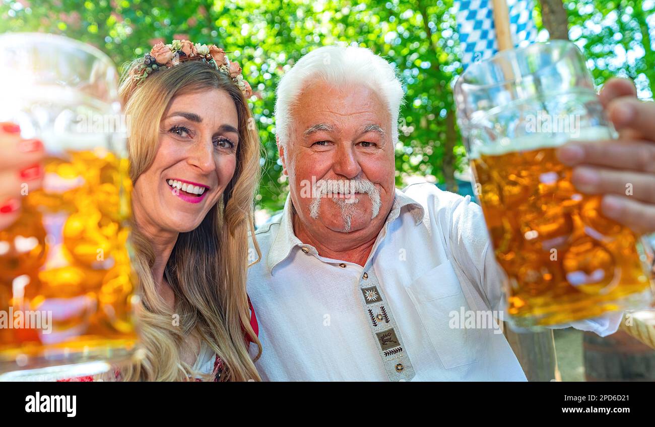 L'uomo anziano e la ragazza giovane che guardano la macchina fotografica dicono i festoni o i bicchieri con la tazza di birra nella birreria all'aperto bavarese o nell'oktoberfest Foto Stock
