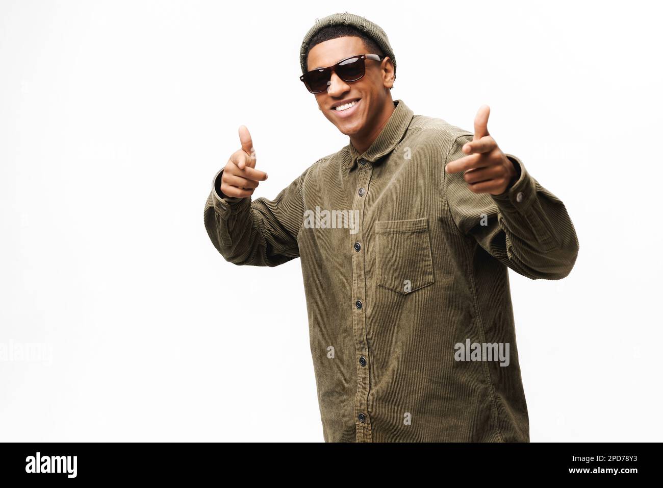 Giocoso afro-americano in occhiali da sole, cappello alla moda e camicia che indica la macchina fotografica isolato su bianco, positivo uomo allegro godendo di divertirsi, scegliendo voi, fingendo di fare un colpo da immaginate pistole Foto Stock