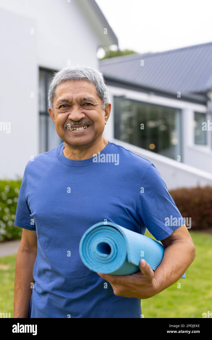 Ritratto di un anziano biraciale sorridente con tappetino yoga in piedi contro casa in cortile Foto Stock