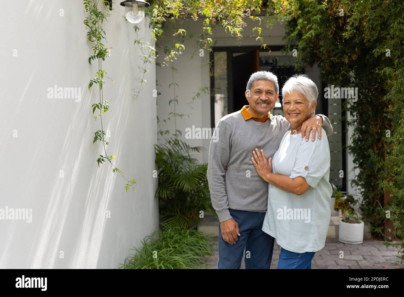 Ritratto di coppia anziana amorevole biraciale sorridente e in piedi da piante in cortile contro casa Foto Stock