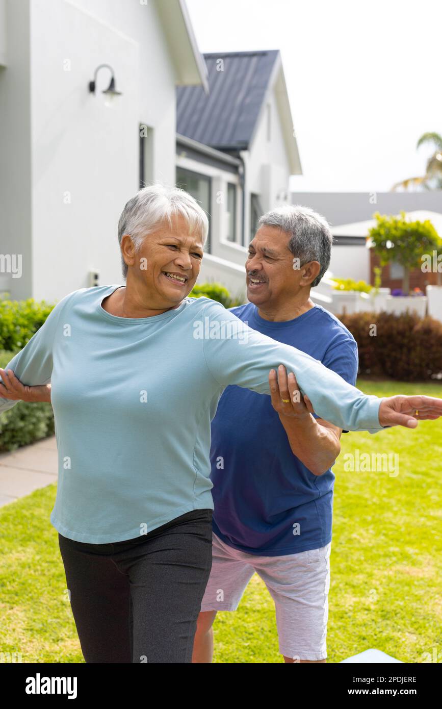 Marito anziano biraciale che assiste la moglie nel praticare la posa guerriero 2 mentre si esercita in cortile Foto Stock