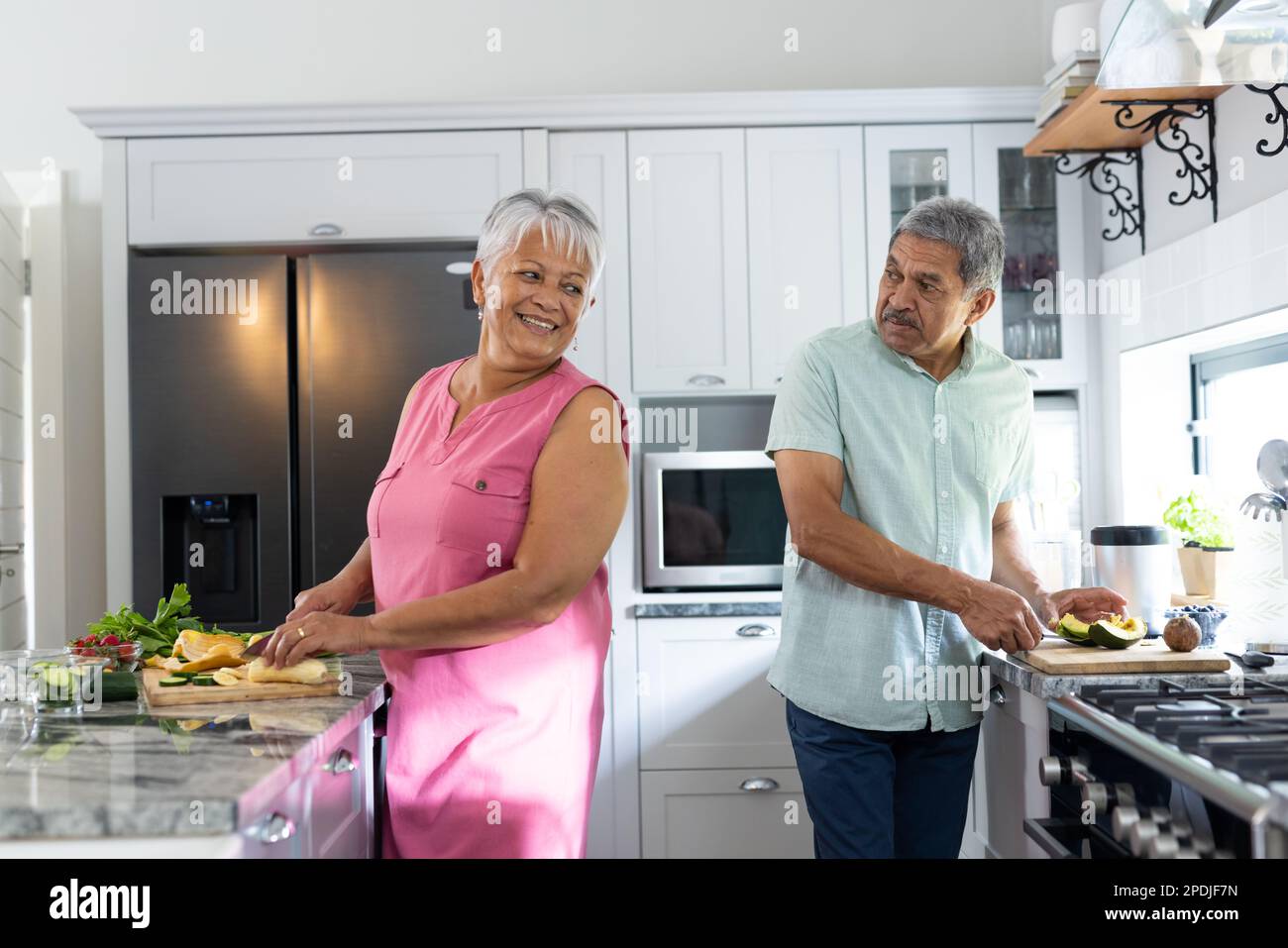 Coppia anziana biraciale che parla e trita le verdure sui banchi della cucina mentre cucinano gli alimenti Foto Stock