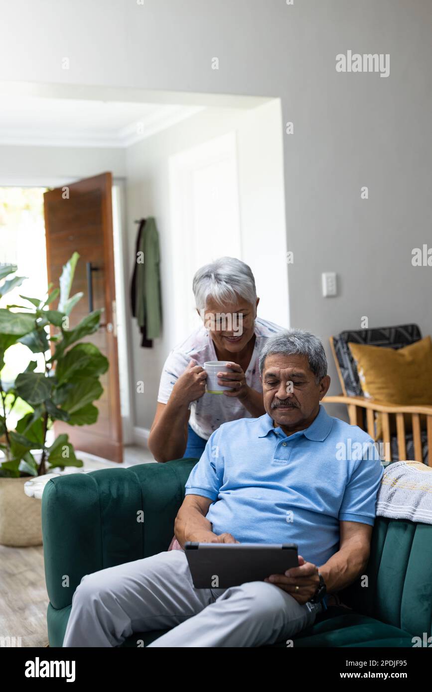 Donna anziana biraciale sorridente con una tazza di caffè appoggiata dal marito utilizzando un tablet digitale sul divano Foto Stock