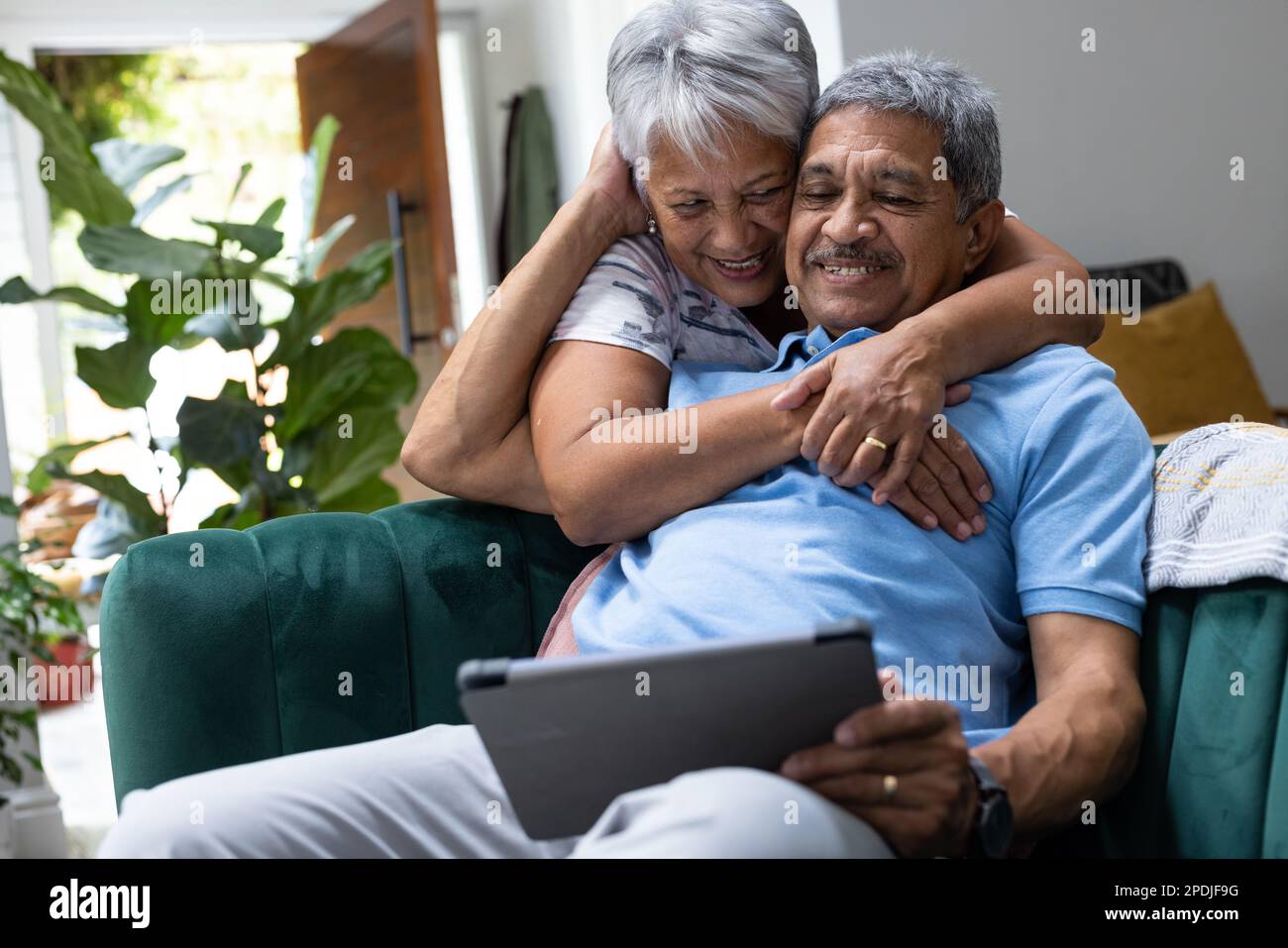 Donna anziana biraciale sorridente che abbraccia il marito da dietro utilizzando un tablet digitale sul divano Foto Stock