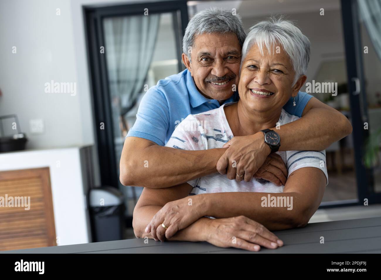 Ritratto di un uomo anziano biraciale sorridente e romantico che abbraccia la moglie da dietro seduta a tavola a casa Foto Stock