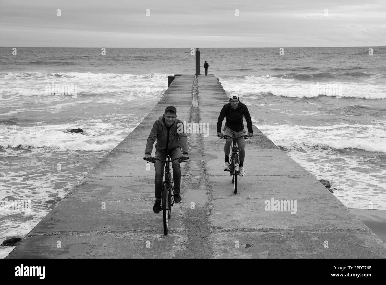 Due ciclisti sul molo con le onde dell'Oceano Atlantico che si infrangono sulla spiaggia, Cascais, Regione di Lisbona, Portogallo, Europa Foto Stock