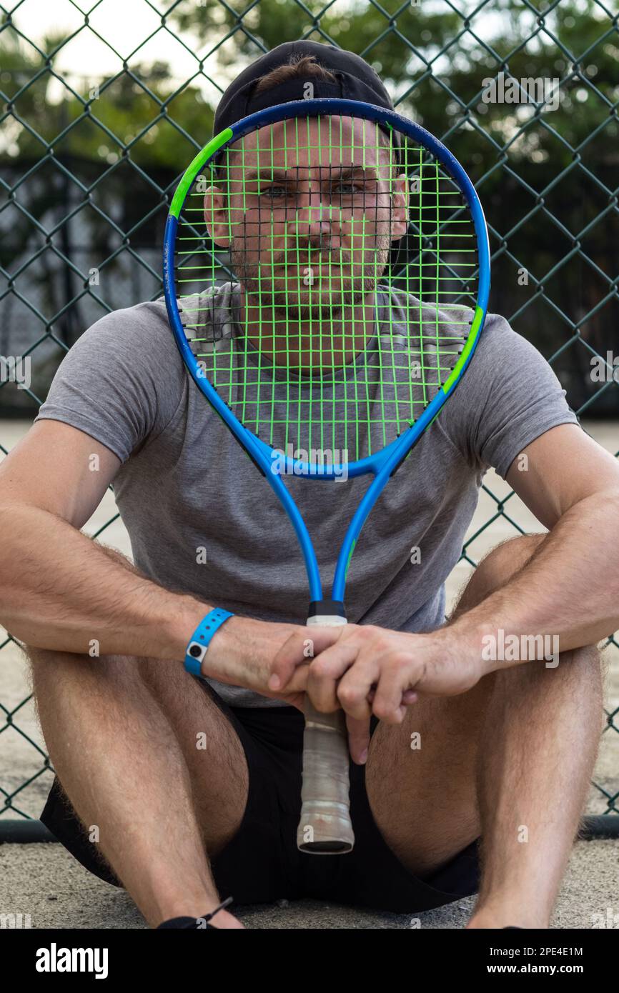 Il giocatore di tennis si siede sul campo da tennis guardando la macchina fotografica attraverso la racchetta da tennis. Foto Stock