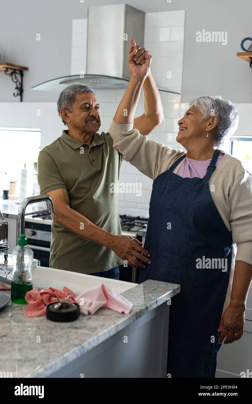 Uomo anziano biraciale che balla con moglie che indossa il grembiule mentre si trova in piedi in cucina, spazio copia Foto Stock