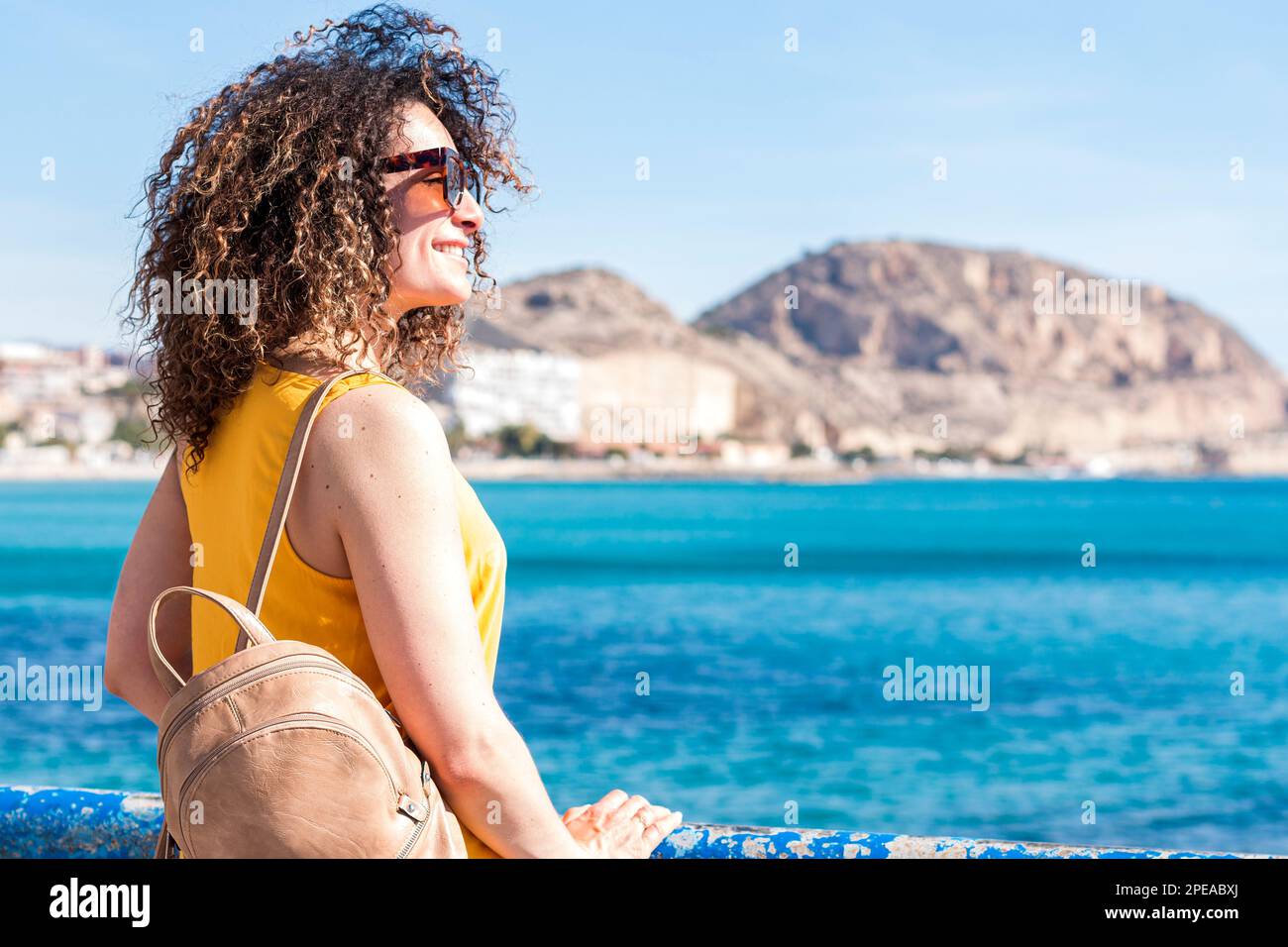 Allegra giovane donna dai capelli ricci in cima giallo e occhiali da sole con zaino in piedi sul molo e sorridente mentre si gode pittoresco mare con mo Foto Stock