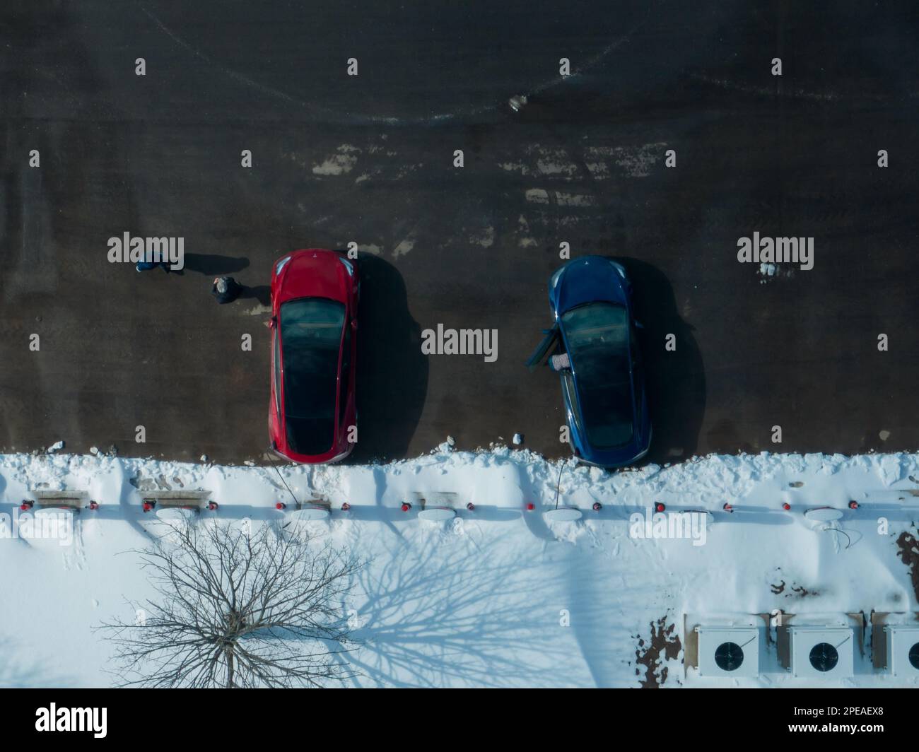 Una foto aerea direttamente sopra due vetture Tesla Model 3 parcheggiate, rossa e blu, presso una stazione Supercharger Tesla nella stagione invernale. Foto Stock