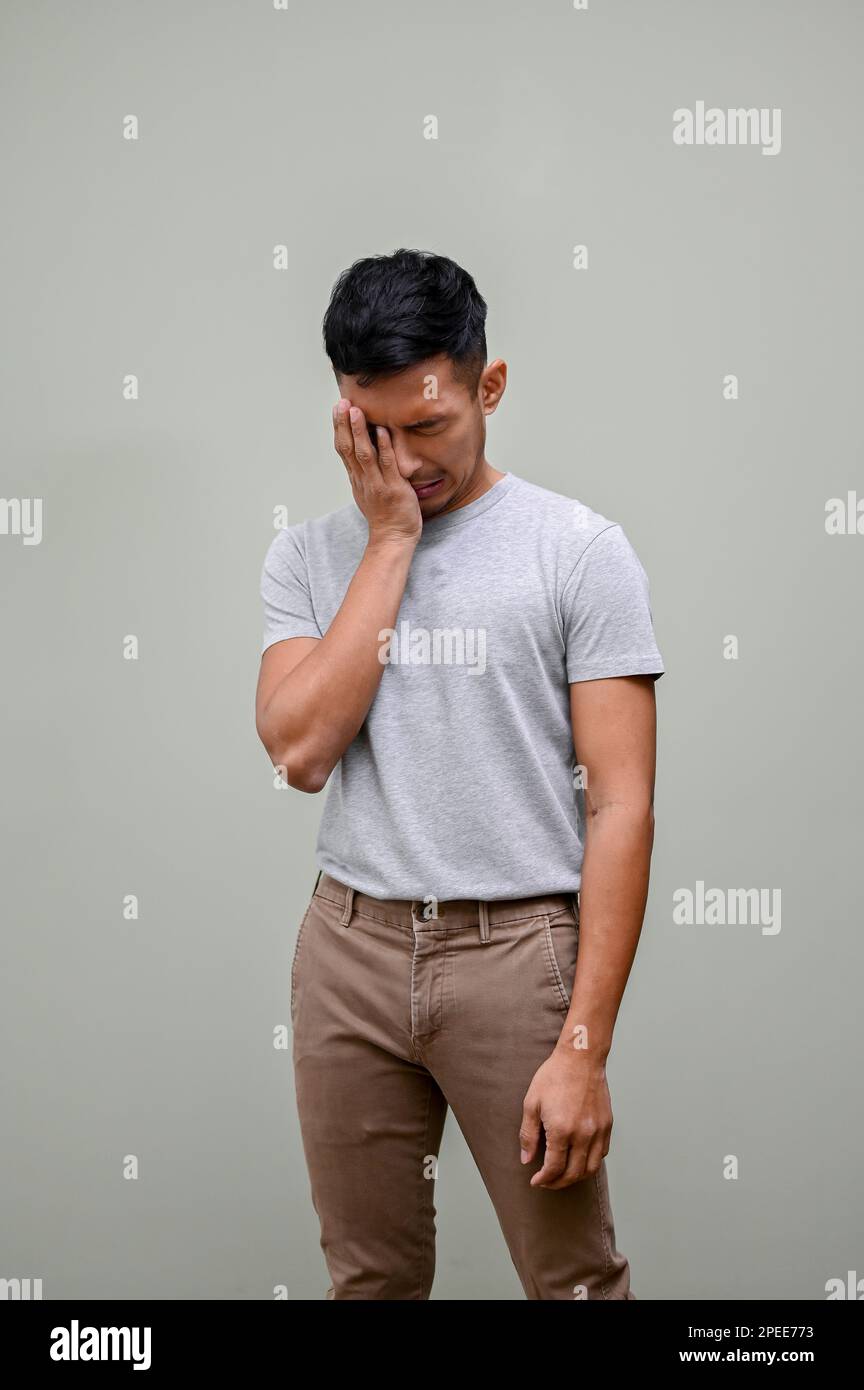 L'uomo asiatico, triste e piangente, in abiti casual che coprono il suo volto, sentendosi sconvolto e fallito, si trova su uno sfondo verde isolato. Foto Stock