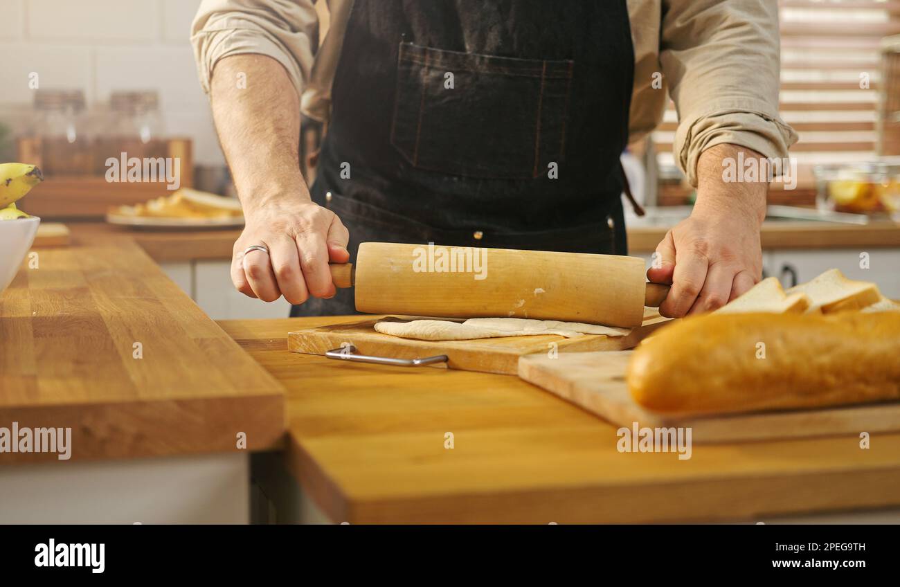 Mani ravvicinate uomo che indossa il grembiule che tiene un matterello, che fa l'impasto sul tavolo da cucina. Foto Stock