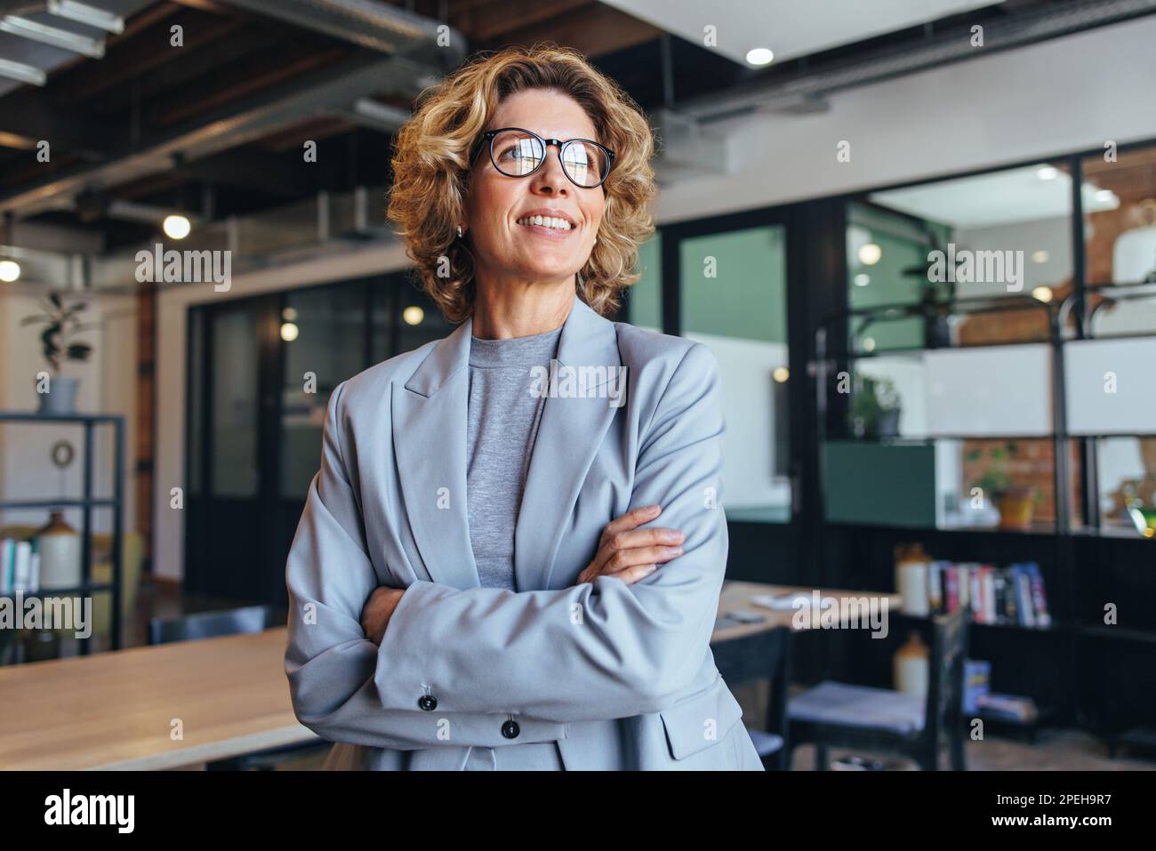 una donna d'affari con i suoi 40 anni guarda lontano con attenzione in un ufficio. Donna professionale in piedi con un vestito con le braccia incrociate. Foto Stock