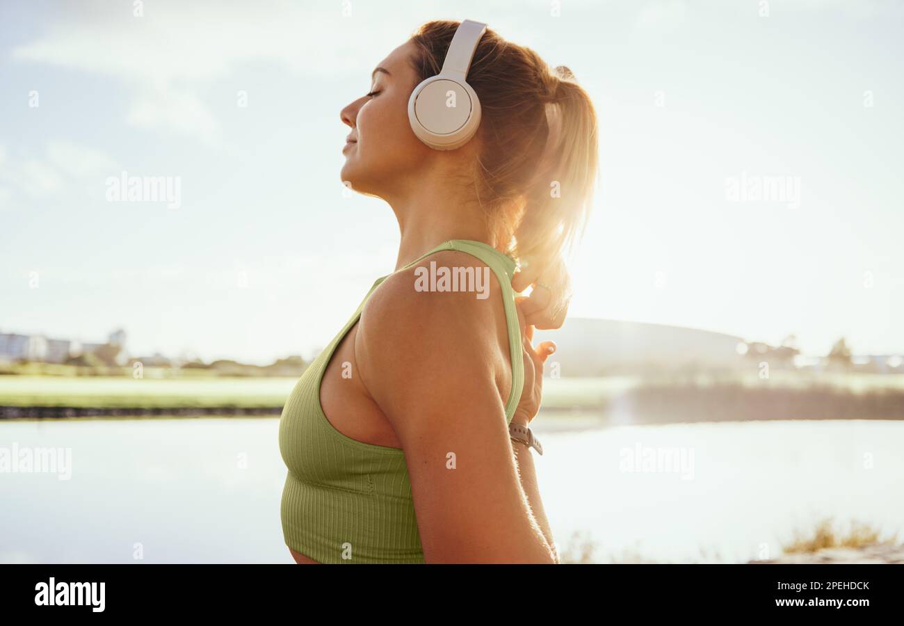 Donna nel suo 30s si prepara ad iniziare la sua routine di esercizio nei pressi di un lago tranquillo. Ha le sue cuffie dentro e sta portando lo sportswear mentre allunga e. Foto Stock