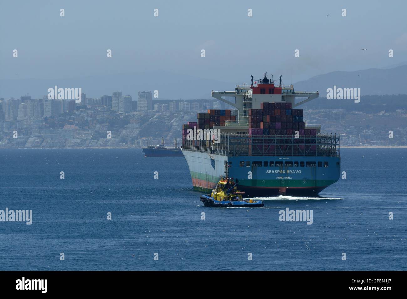 Porto di Valparaiso - Container Freighter in partenza dal porto accompagnato da un rimorchiatore giallo e blu. Acque calme con Valparaiso sullo sfondo. Foto Stock