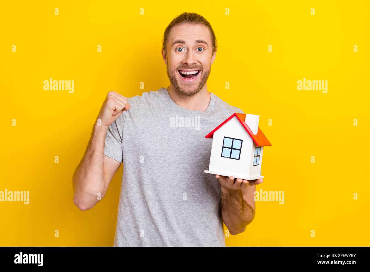 Foto di uomo fortunato positivo indossare t-shirt grigia che tiene piccola casa nascente pugno isolato sfondo di colore giallo Foto Stock