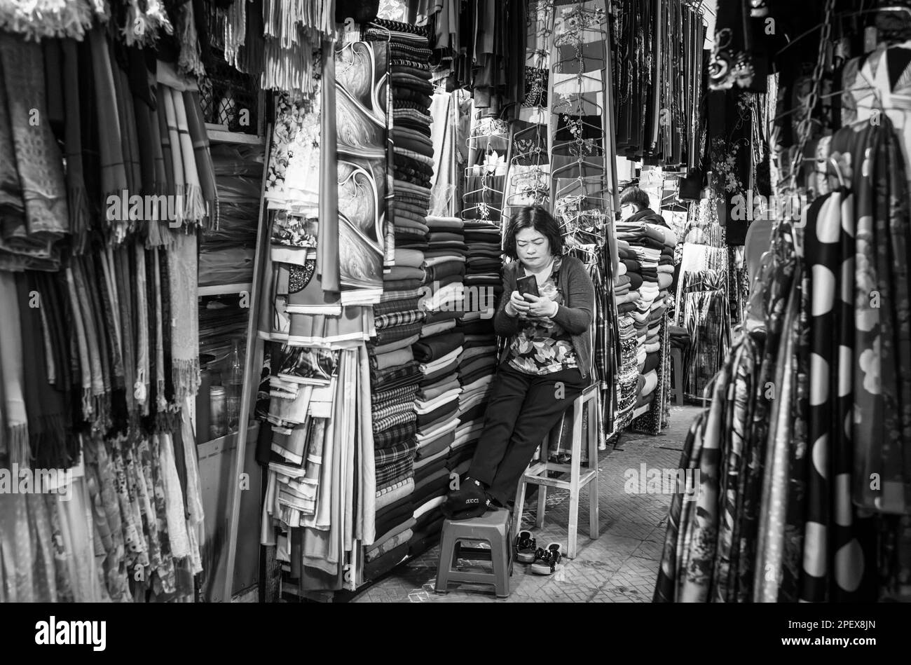Una donna vietnamita stalla porta guarda il suo cellulare come lei attende i clienti al suo tessuto stand a Dalat Market, Vietnam. Foto Stock