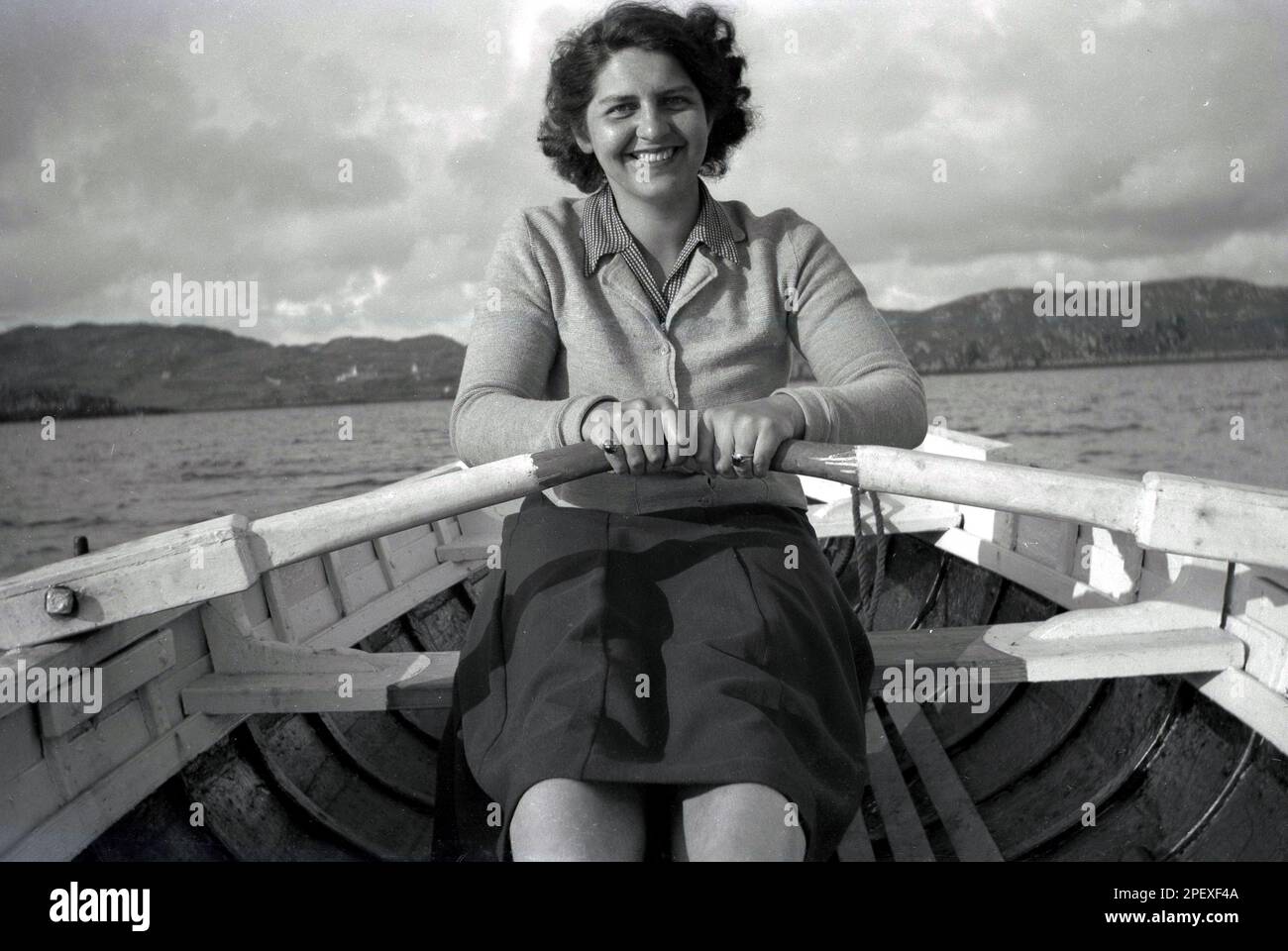 1950s, storica, fuori sul mare sulla costa, una giovane donna, in cima e gonna, in vacanza, divertendosi a remi su una barca di legno, Inghilterra, Regno Unito. Foto Stock