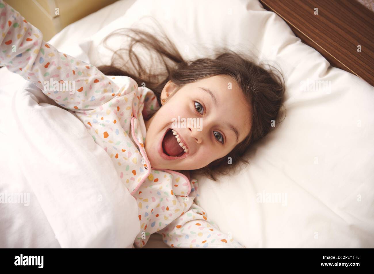 Bambina maliziosa in pigiama, che fa volti alla macchina fotografica, che si allunga svegliandosi al mattino, rilassandosi nel suo letto Foto Stock