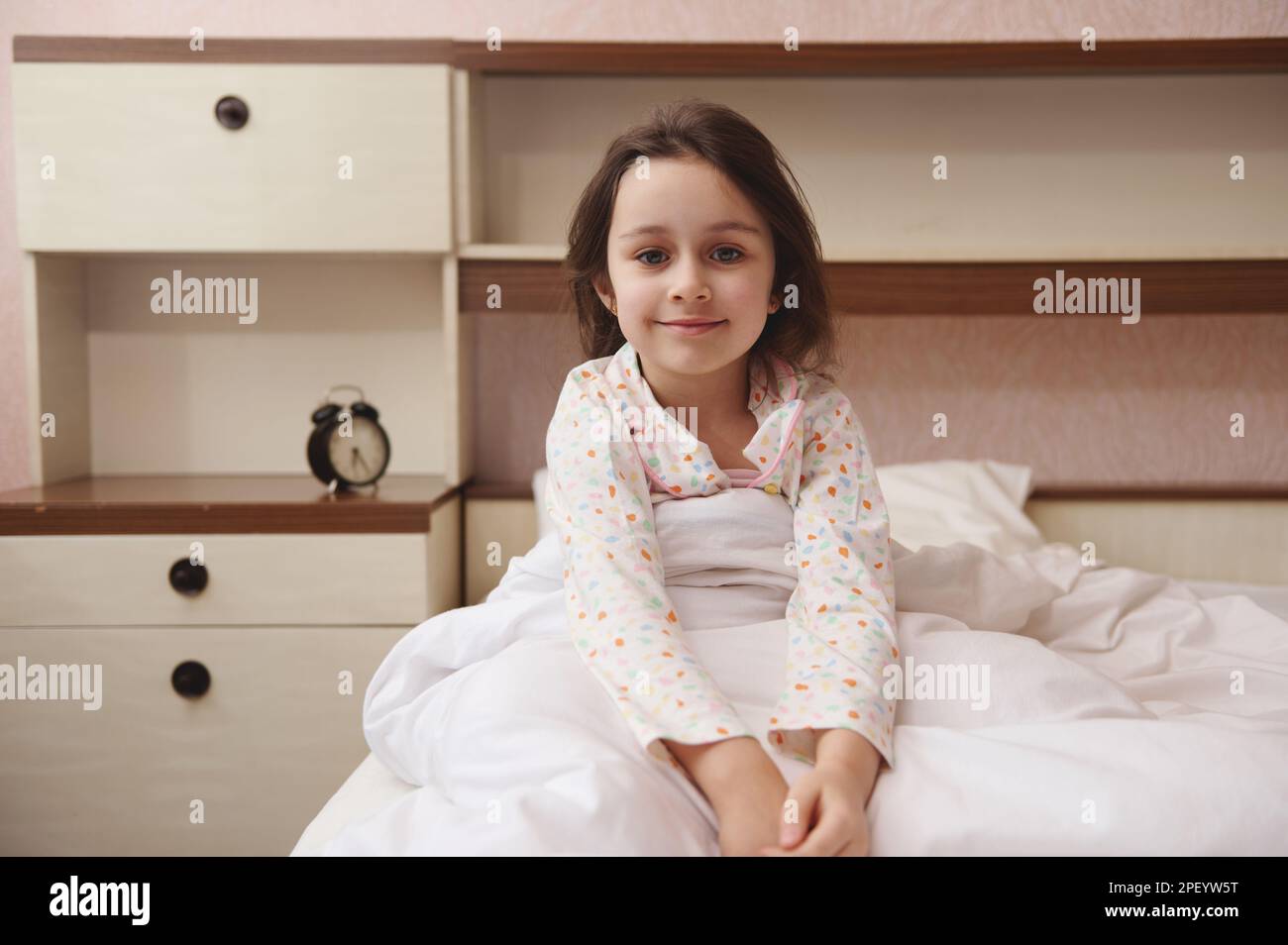Bella bambina in pigiama, sorridente guardando la macchina fotografica, seduta a letto nella sua camera da letto accogliente. Bambini felici Foto Stock