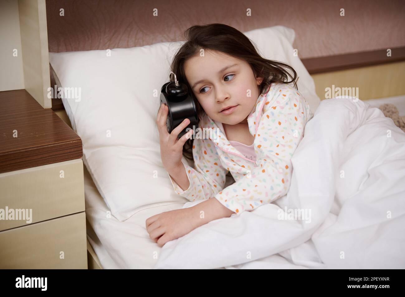 Adorabile bambina che tiene una sveglia vicino al suo viso, vestita di pigiami glamour, preparandosi per il letto Foto Stock